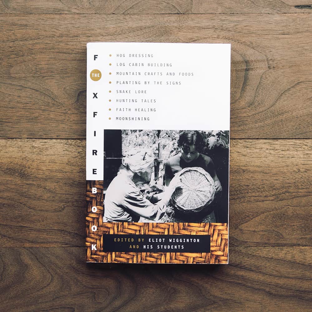 Cover of foxfire book against a wooden table. Cover shows older woman helping younger studen make a basket